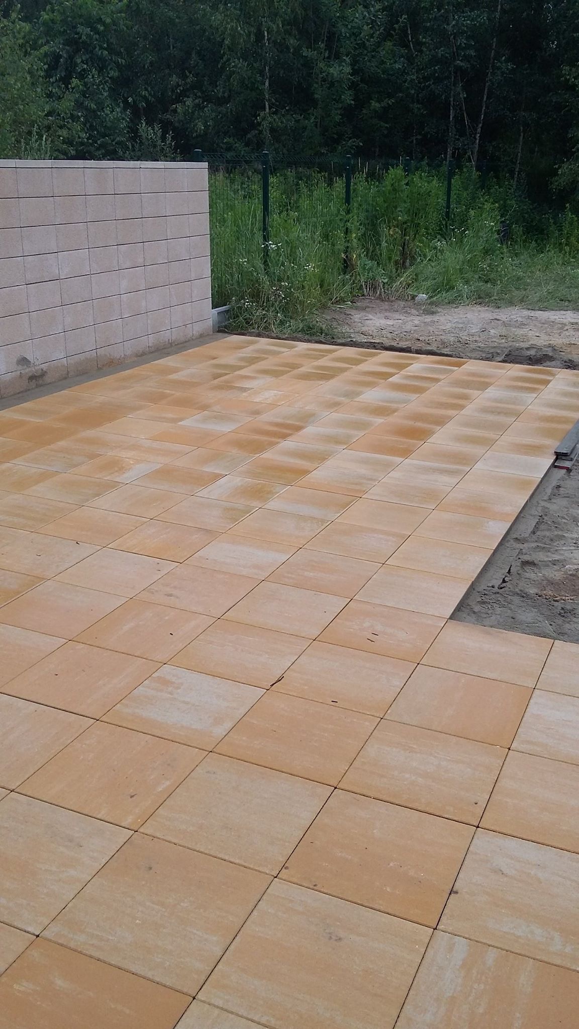 Żółta kostka brukowa ułożona na posesji, widoczny fragment muru z bloczków i trawa za ogrodzeniem. Częściowo ukończona realizacja.