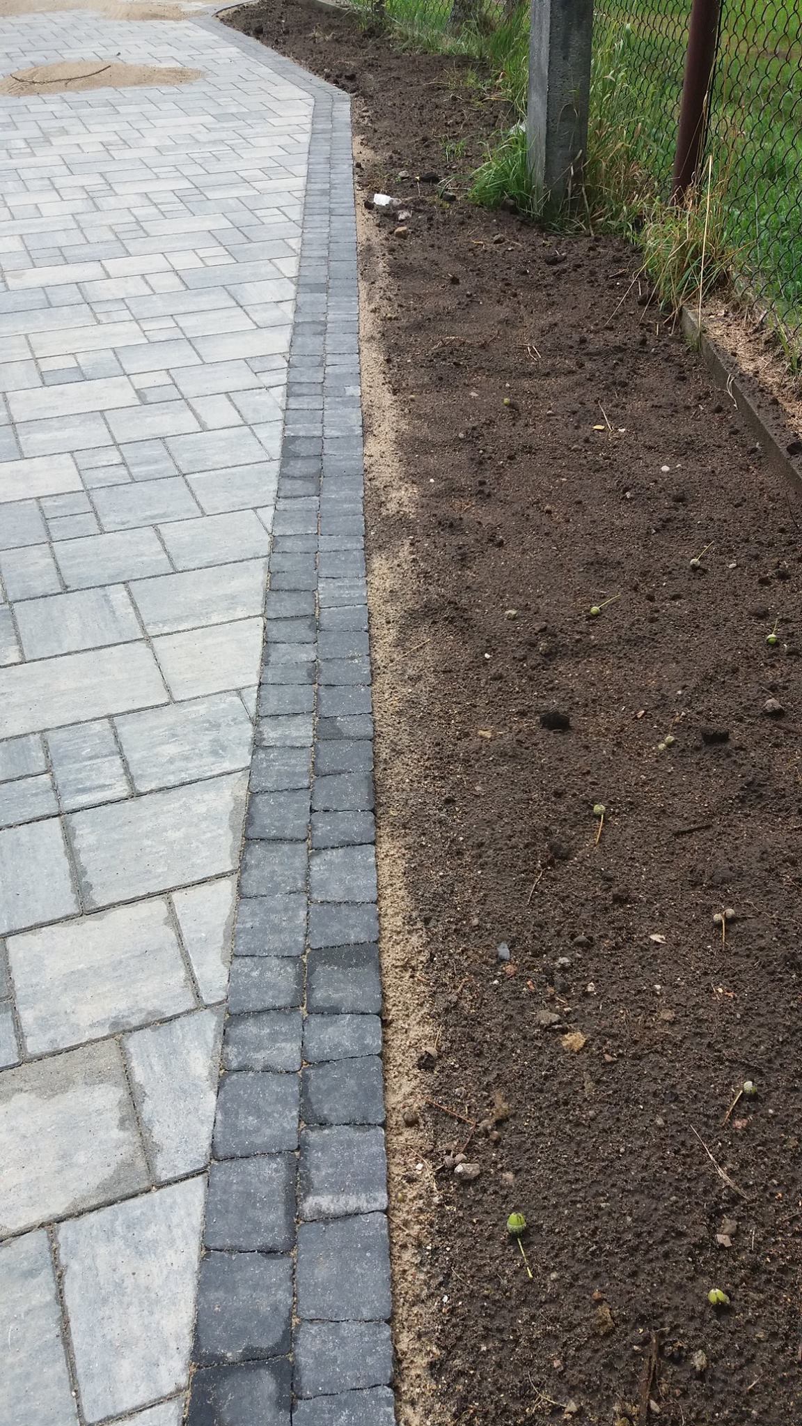Szara kostka brukowa ułożona wzdłuż krawędzi z ciemniejszą obwódką, przylegająca do ziemi z widocznymi fragmentami roślinności i siatki ogrodzeniowej.