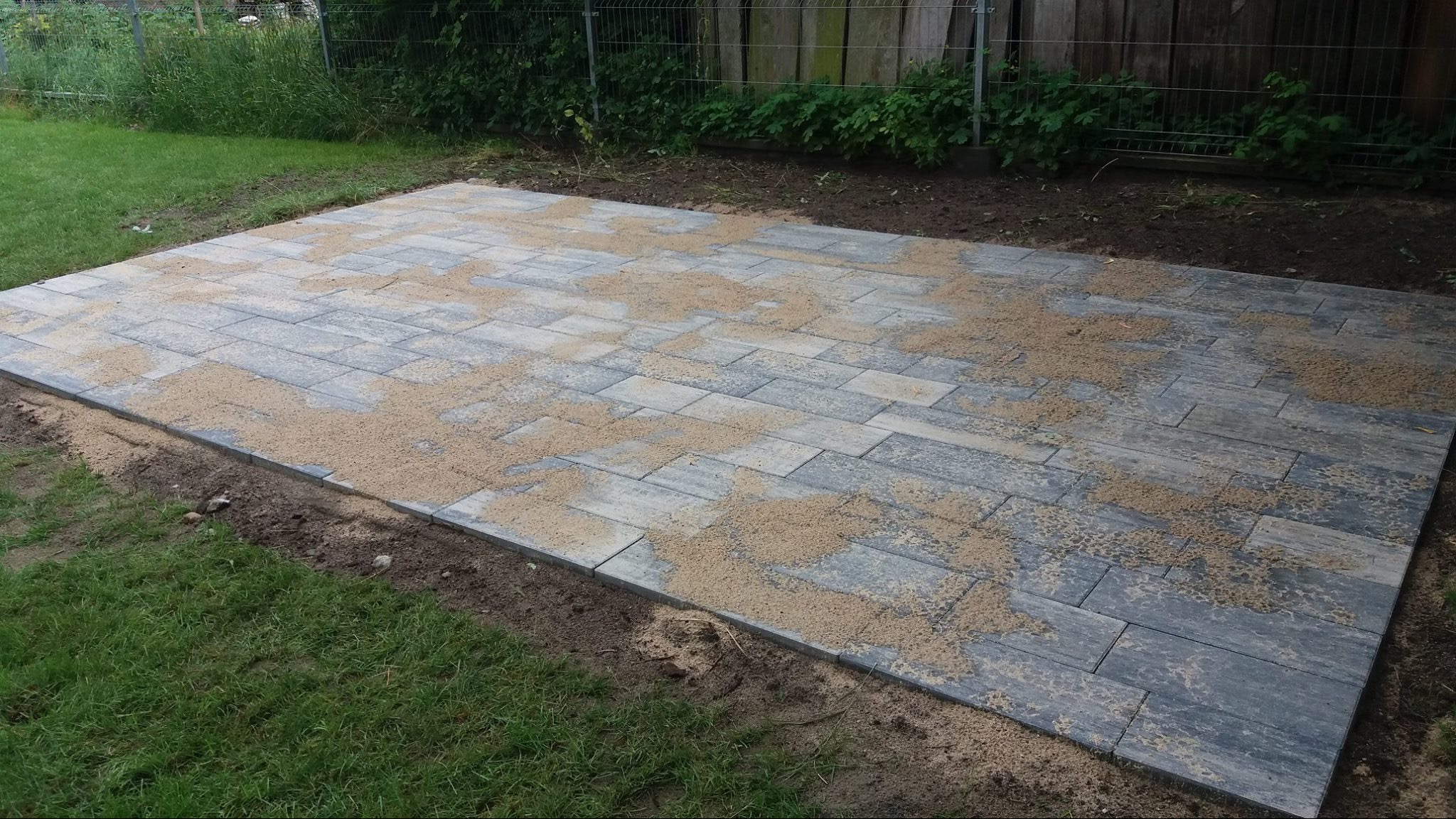Ułożona, prostokątna nawierzchnia z szarej kostki brukowej imitującej drewno, zasypana piaskiem, na tle trawnika i ogrodzenia z siatki.