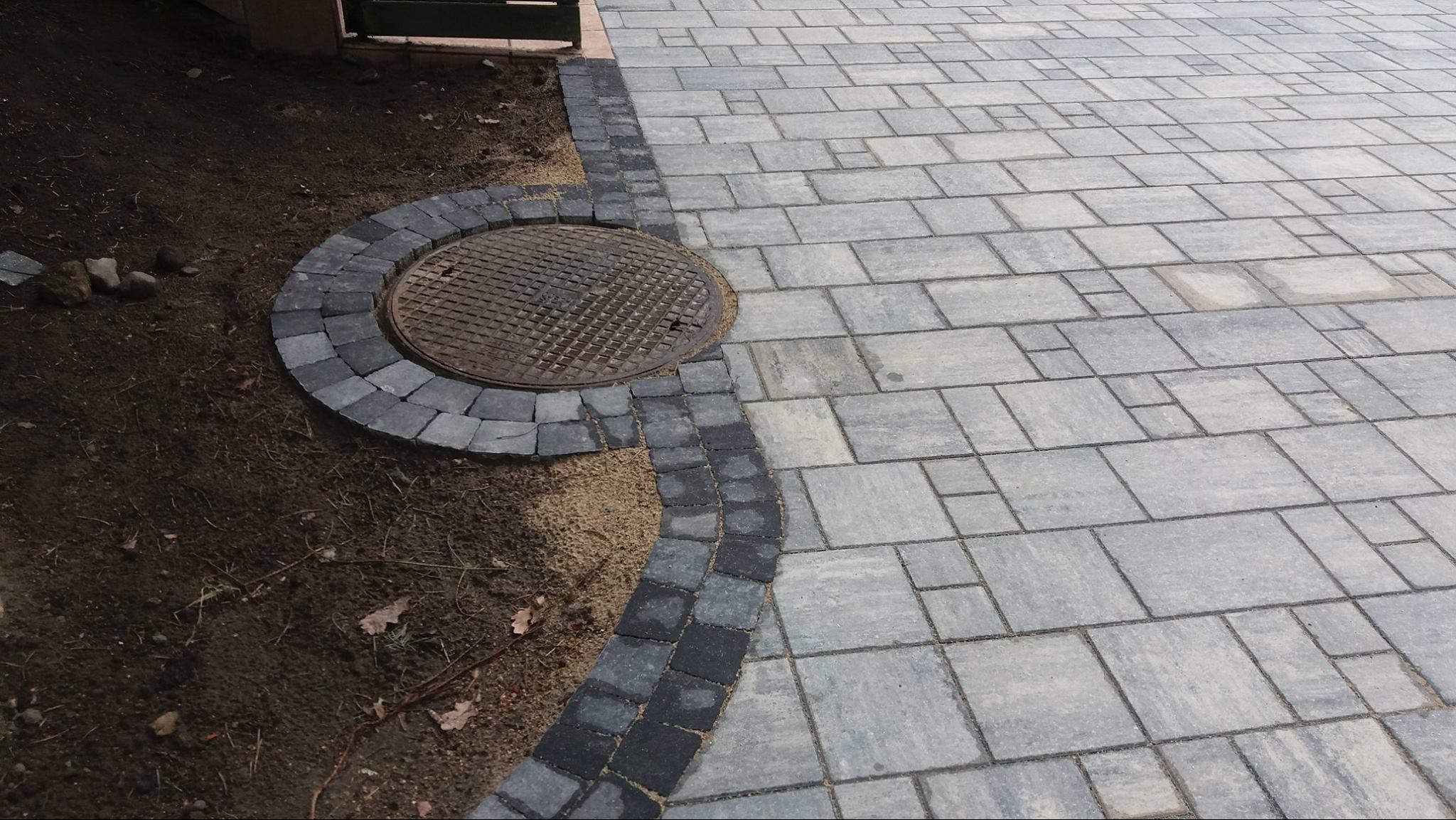 Ułożona szara kostka brukowa z ciemną obwódką i studzienką kanalizacyjną. W tle ziemia i fragment ogrodzenia. Regularny wzór kostki.