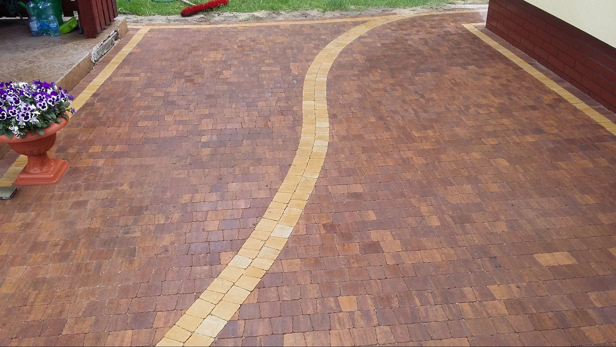 Brązowa kostka brukowa z żółtą obwódką i falującym pasem, donica z kwiatami w lewym rogu, widok z góry.