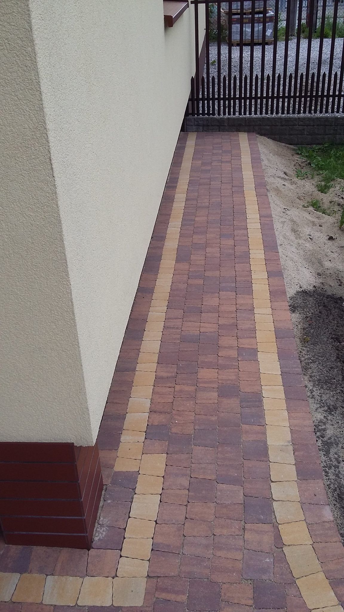 Ułożony chodnik z kostki brukowej w odcieniach brązu i beżu, prowadzący wzdłuż ściany budynku z widocznym fragmentem ogrodzenia w tle.