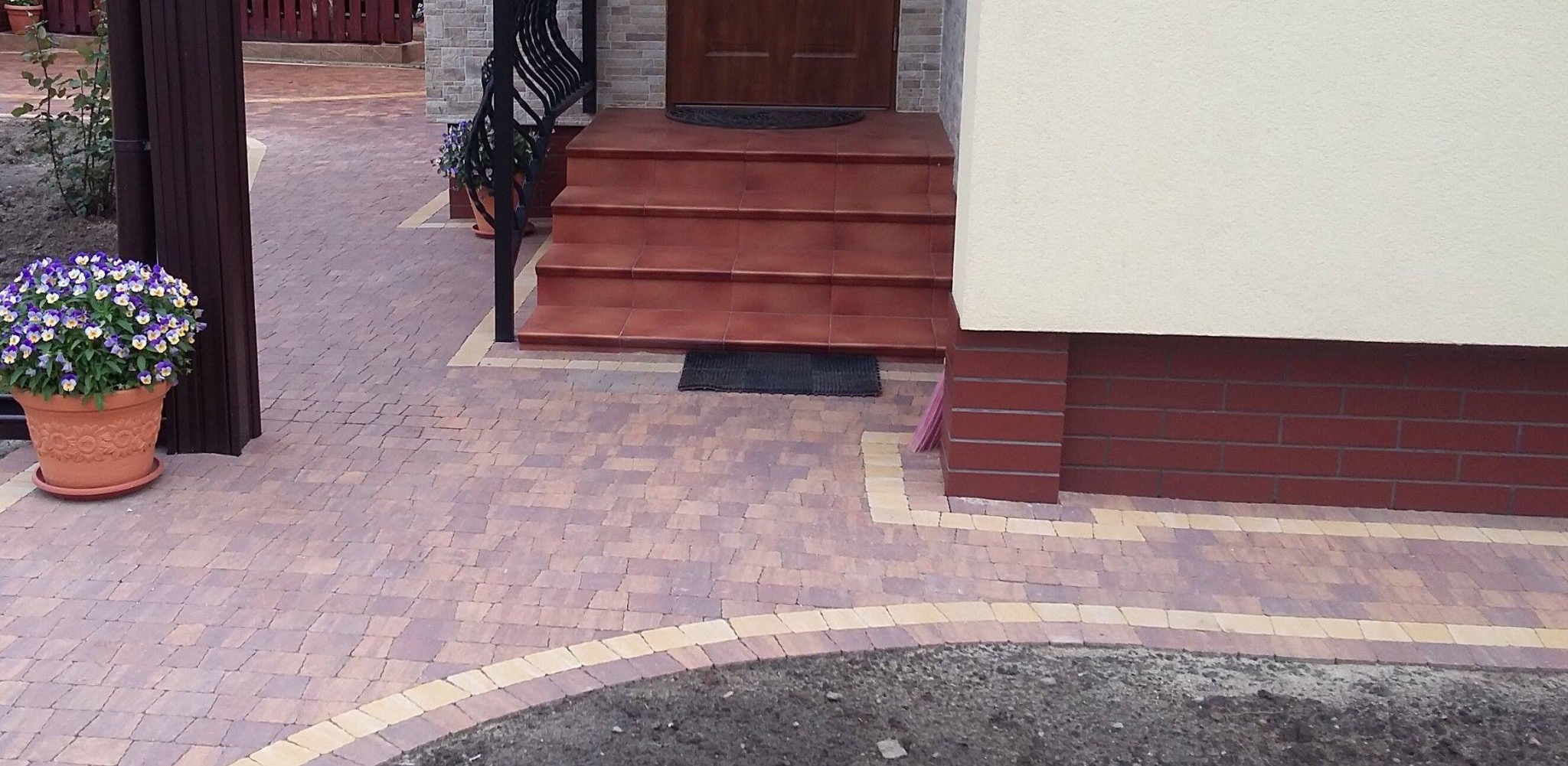 Ułożona kostka brukowa w odcieniach brązu i beżu tworzy wzór na podjeździe. Schody wejściowe z brązowych płytek, donica z fioletowymi bratkami.