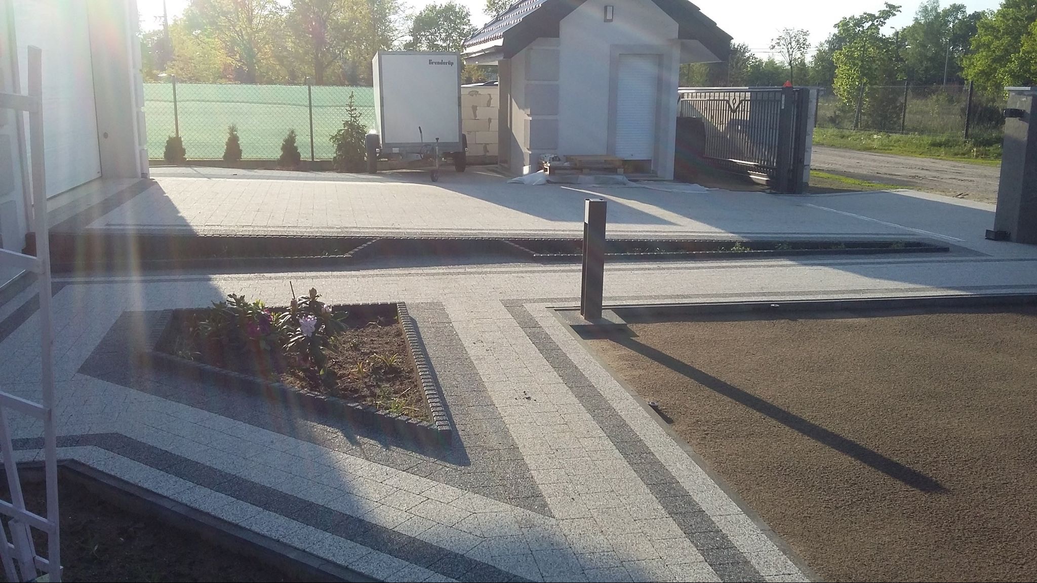 Ułożona kostka brukowa w dwóch odcieniach szarości tworząca wzór na podjeździe. Widoczny fragment ogrodu i budynek gospodarczy.