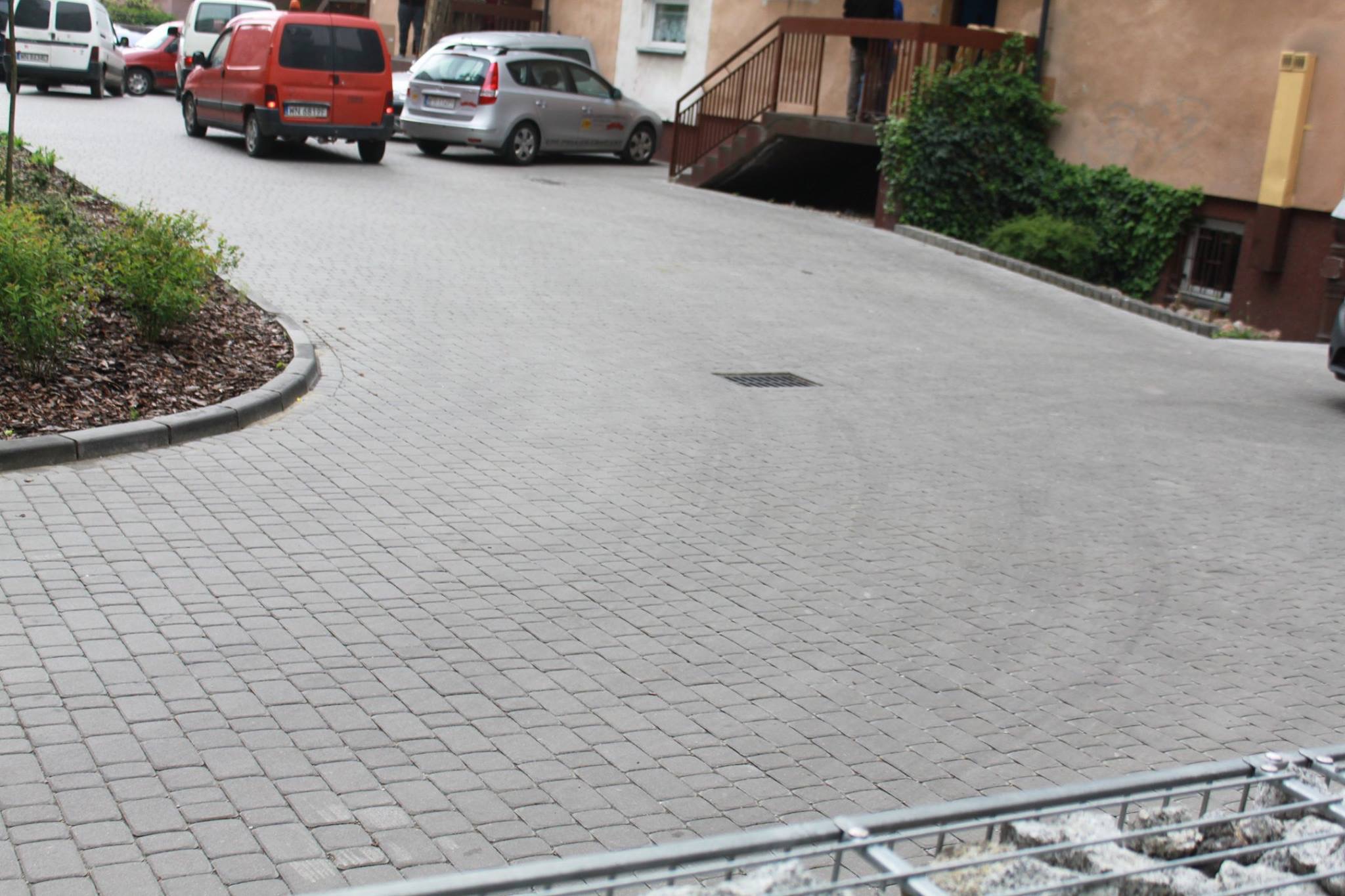 Szara kostka brukowa ułożona na podjeździe, z widoczną krawędzią trawnika obsadzoną roślinami. W tle zaparkowane samochody i budynek z wejściem do piwnicy.