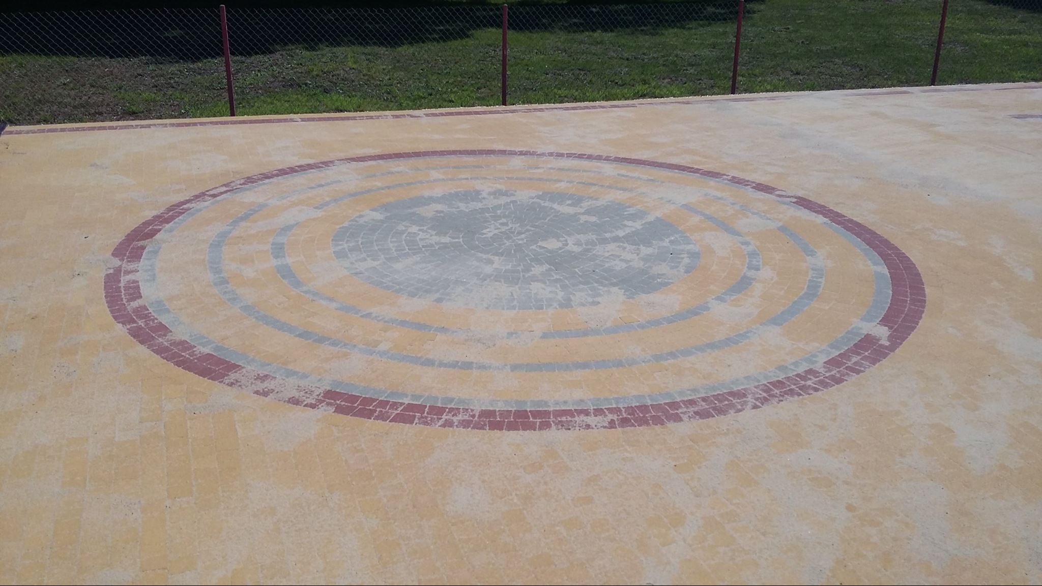Okrąg z kostki brukowej w odcieniach szarości, beżu i czerwieni, tworzący koncentryczne kręgi na placu. Widoczne ogrodzenie w tle.
