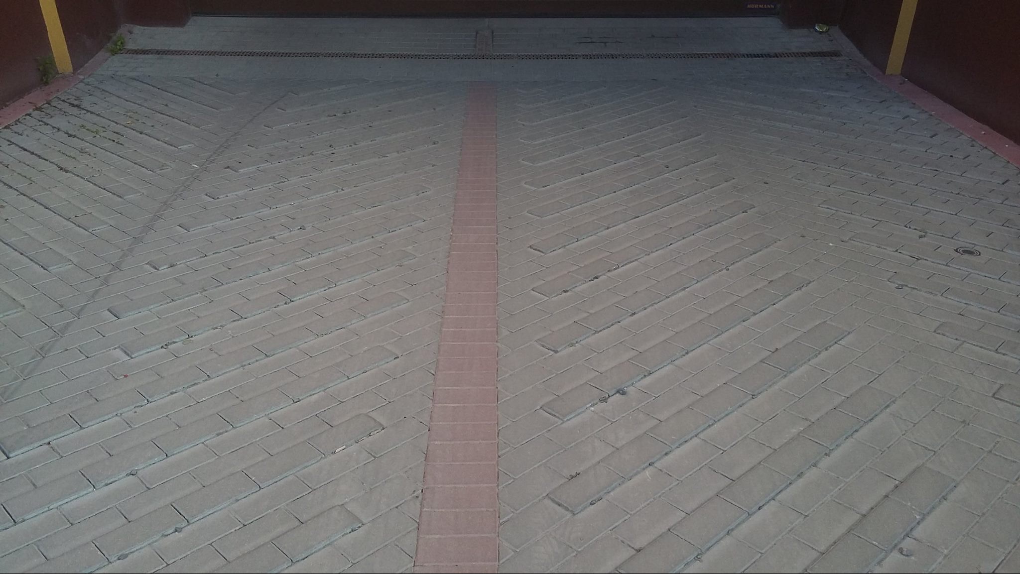 Ułożona kostka brukowa w jodełkę z centralnym pasem z czerwonej kostki. Widoczny odpływ liniowy. Brama garażowa w tle.