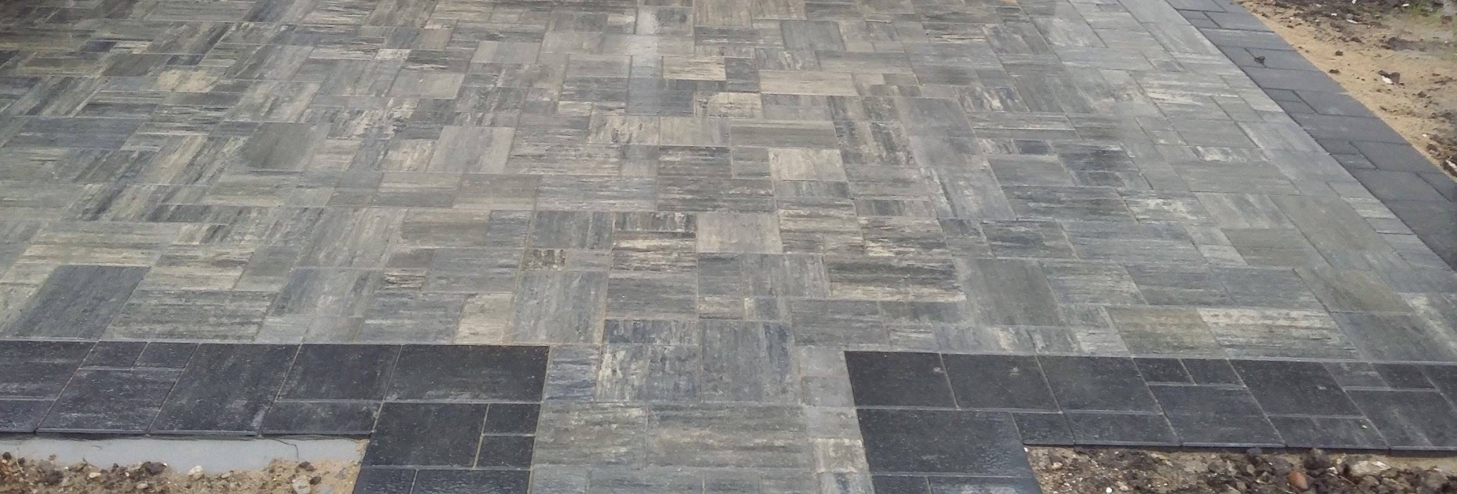 Ułożona kostka brukowa w odcieniach szarości, z ciemniejszym obramowaniem. Widoczny fragment niedokończonej krawędzi.