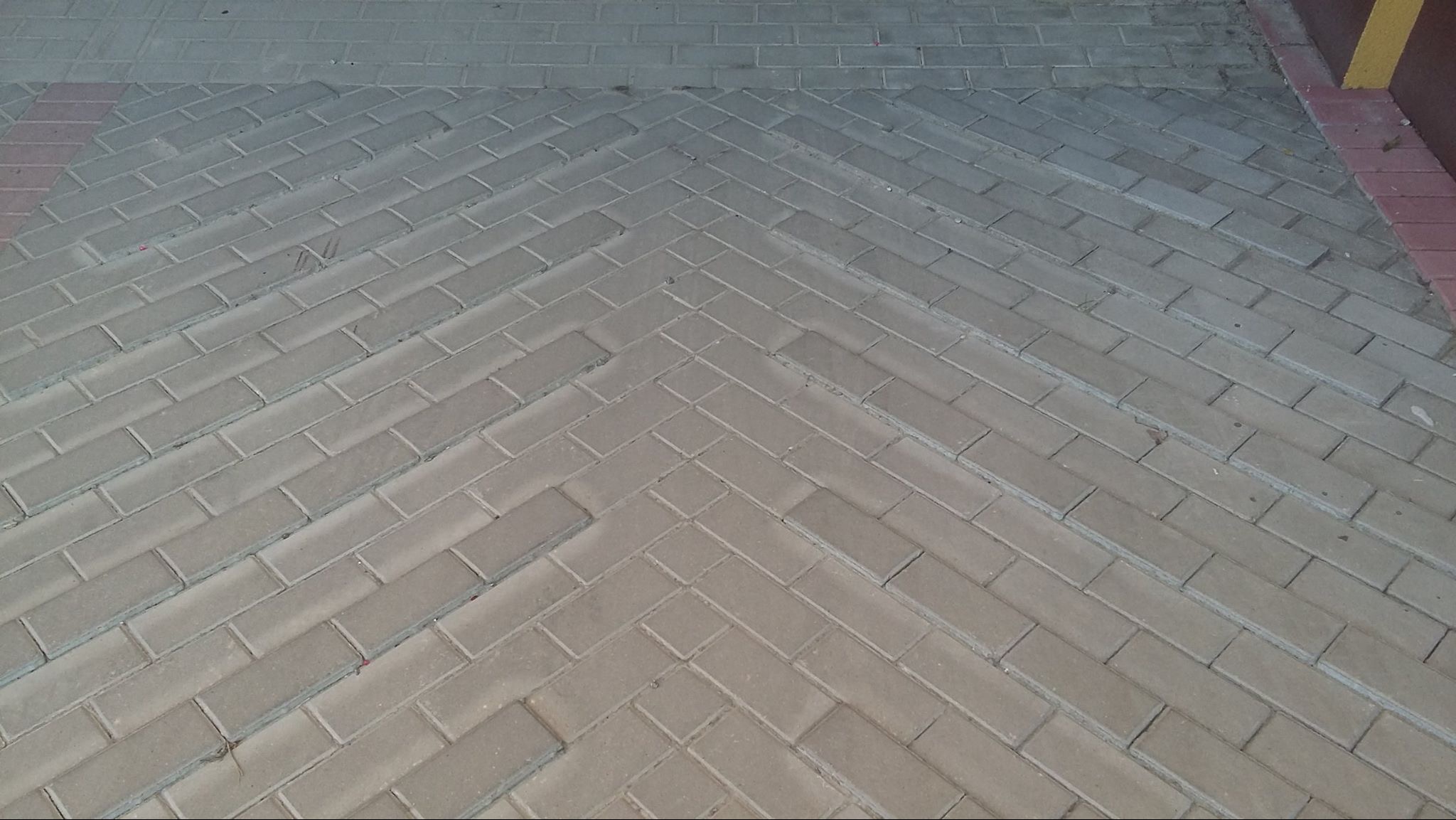 Ułożona, szara kostka brukowa w jodełkę, z delikatnym cieniowaniem i czerwoną obwódką przy ścianie. Widok z góry, brak osób.