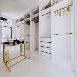 Vastriya Design - Nowoczesna garderoba w bieli, z złotymi akcentami. Widoczny stół z dekoracjami, w tle półki z ubraniami i akcesoriami. Marmurowa podłoga dodaje elegancji.