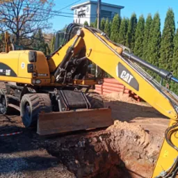 Żółta koparko-ładowarka CAT na tle wykopu i zielonych tuj. Widoczne rury z tworzywa, taśmy ostrzegawcze i hydrant. W tle budynek i drzewa.