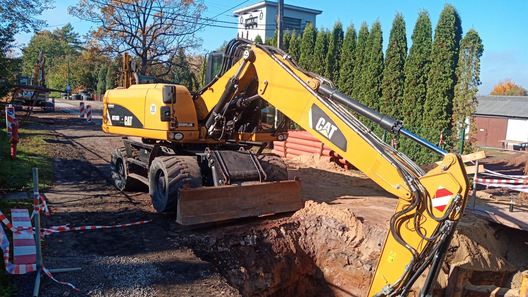 Żółta koparko-ładowarka CAT na tle wykopu i zielonych tuj. Widoczne rury z tworzywa, taśmy ostrzegawcze i hydrant. W tle budynek i drzewa.