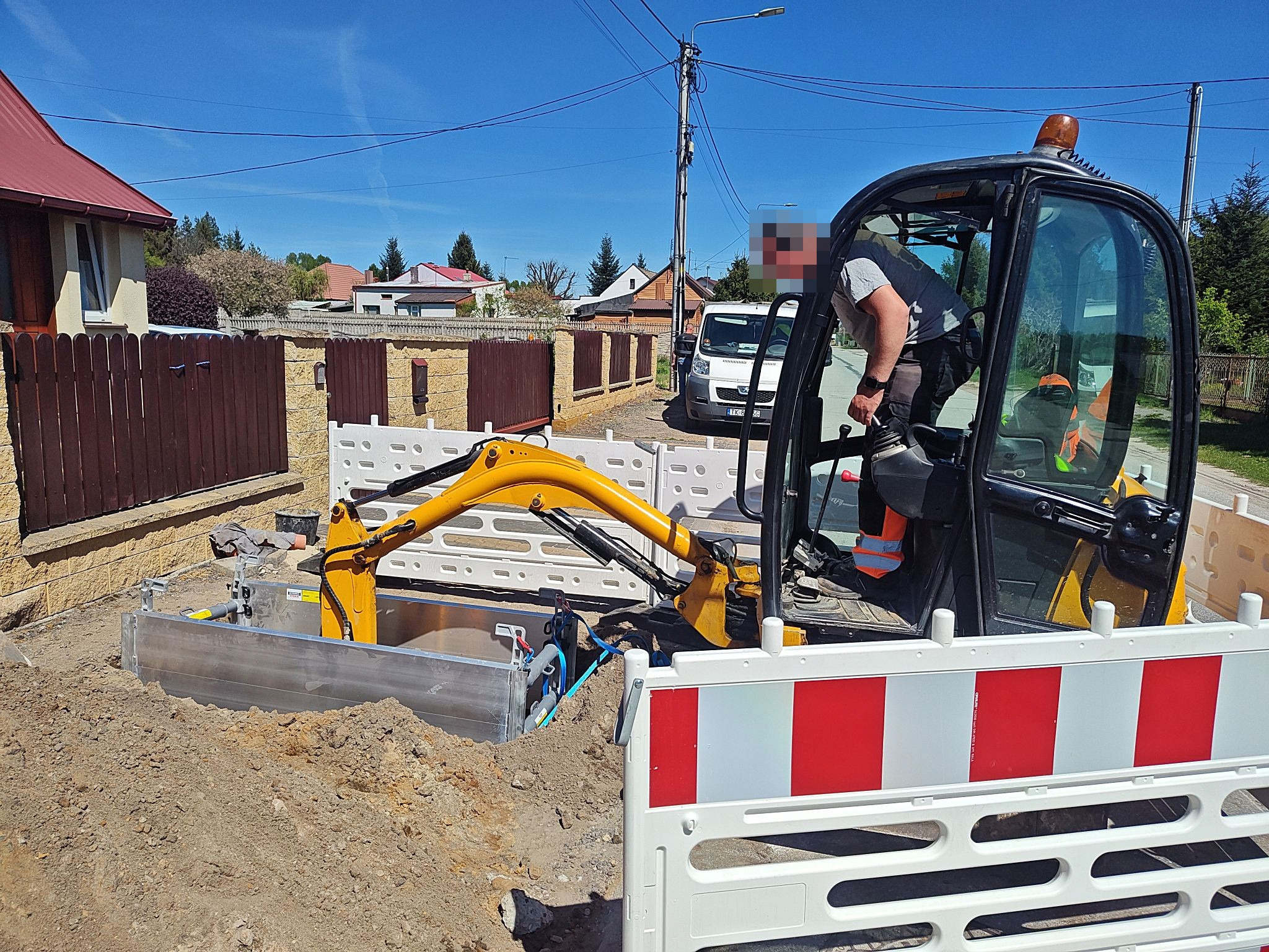 Żółta mini koparka w wykopie zabezpieczonym metalową szalunką i barierkami. Operator w kabinie, w tle domy i błękitne niebo. Prace ziemne w słoneczny dzień.