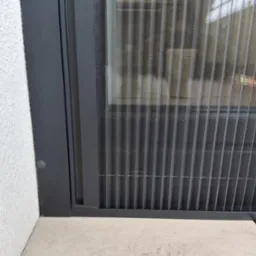 Zamknięta, szara moskitiera plisowana w oknie, widok z bliska na strukturę siatki i ramę, z fragmentem białej ściany z fakturą i jasną posadzką w tle.