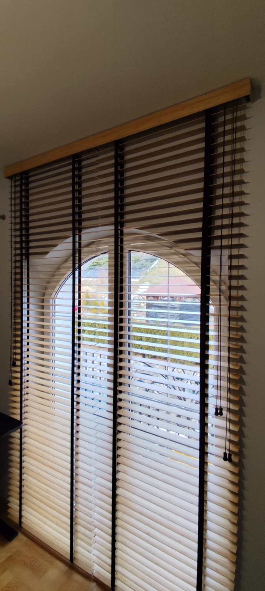 Jasne żaluzje drewniane z czarnymi taśmami na oknach o łukowatym kształcie, widok na zewnątrz z zielenią i zabudowaniami. U góry drewniana listwa mocująca.