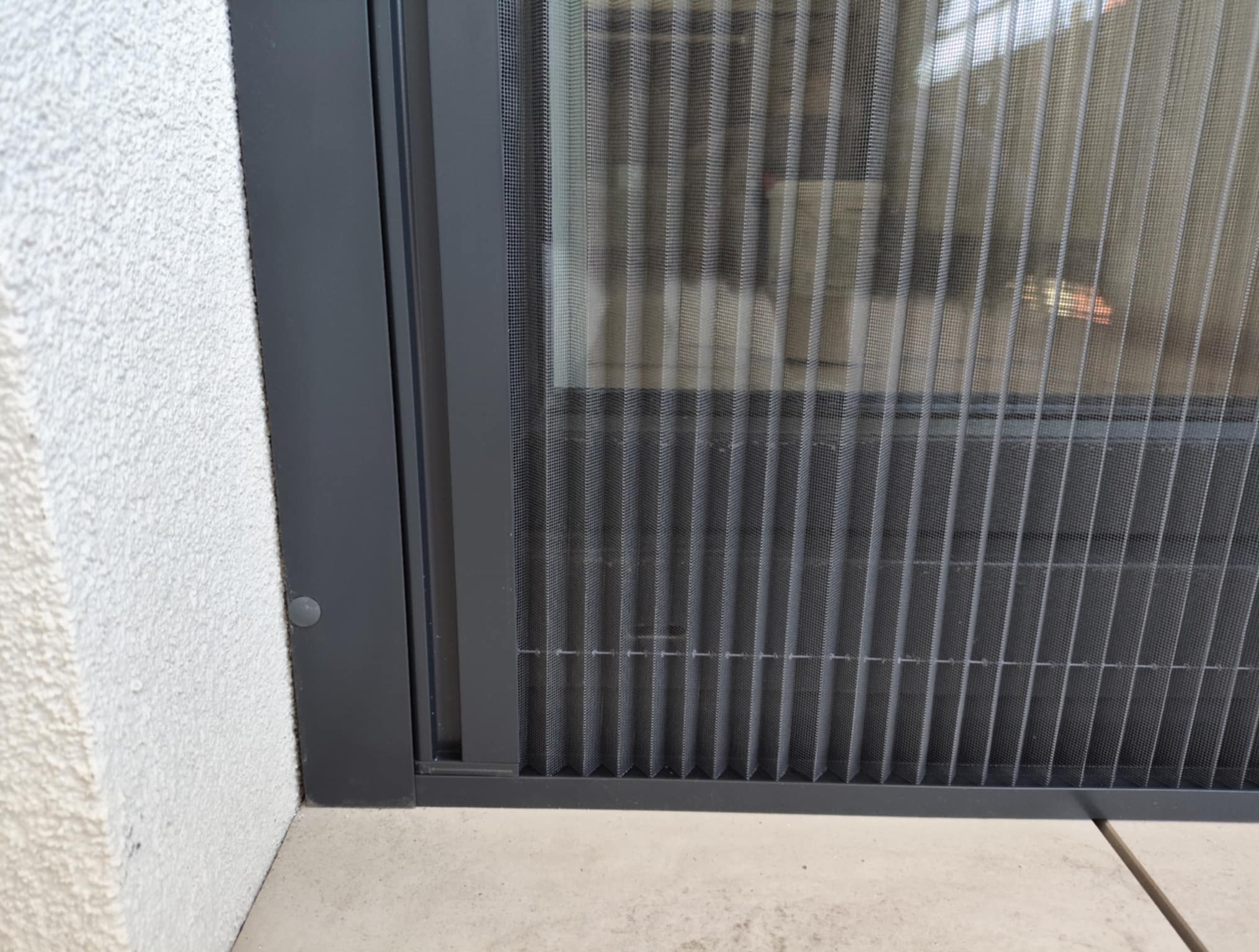 Zamknięta, szara moskitiera plisowana w oknie, widok z bliska na strukturę siatki i ramę, z fragmentem białej ściany z fakturą i jasną posadzką w tle.