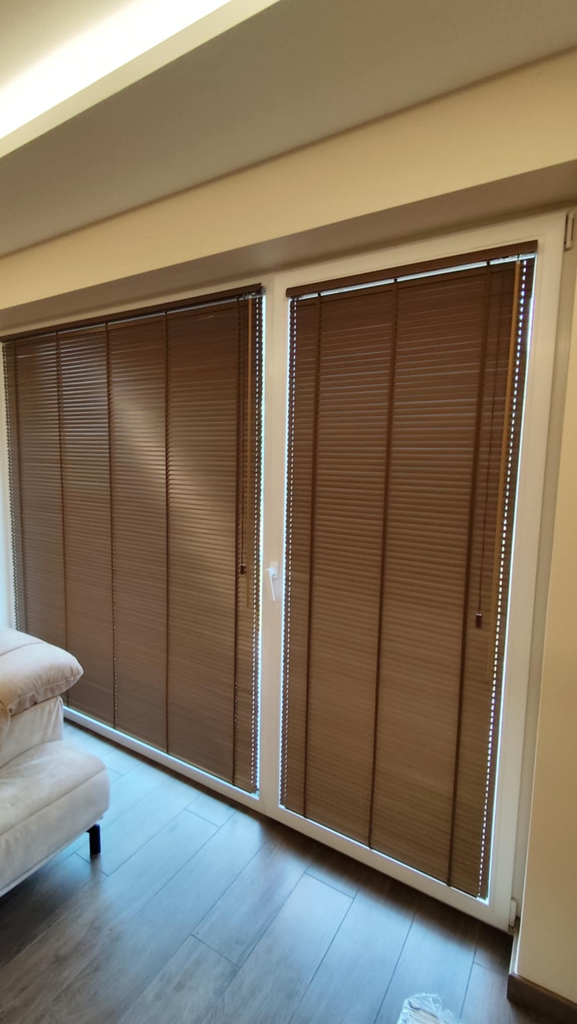 Brązowe plisy okienne zamontowane na dwuskrzydłowym oknie balkonowym z białymi ramami, widok z wnętrza pomieszczenia.