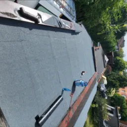 Nowe pokrycie dachu z papy termozgrzewalnej na budynku jednorodzinnym. Widoczne narzędzia dekarskie i butla gazowa. Tło stanowią drzewa i zabudowa mieszkalna.