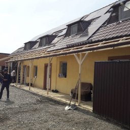 Dachy i rynny - Montaż drewnianej konstrukcji zadaszenia przy żółtym budynku z brązowym dachem i panelami słonecznymi. Widoczni pracownicy, narzędzia i gruz na ziemi.