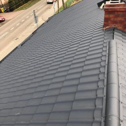 Dachy i rynny - Nowy, grafitowy dach z blachy trapezowej w Katowicach. Widoczny fragment ulicy i komin. Fachowiec na dachu podczas prac dekarskich.