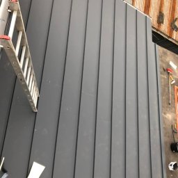 Dachy i rynny - Szary dach z blachy trapezowej z drabiną opartą o jego krawędź. Widoczne narzędzia i materiały dekarskie. Dach sąsiaduje z rdzewiejącym dachem z blachy falistej.