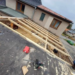 Dachy i rynny - Demontaż starego dachu z widoczną konstrukcją więźby, resztki pokrycia na membranie, narzędzia (piła, kanister) i fragment budynku w tle. Ujęcie z góry.