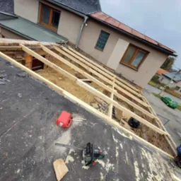 Demontaż starego dachu z widoczną konstrukcją więźby, resztki pokrycia na membranie, narzędzia (piła, kanister) i fragment budynku w tle. Ujęcie z góry.