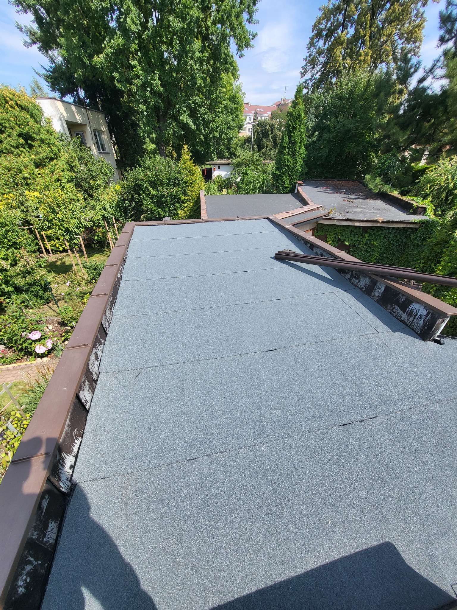 Szary, płaski dach pokryty papą termozgrzewalną, z widoczną obróbką blacharską i zielonym otoczeniem. Widok z góry na fragment dachu.