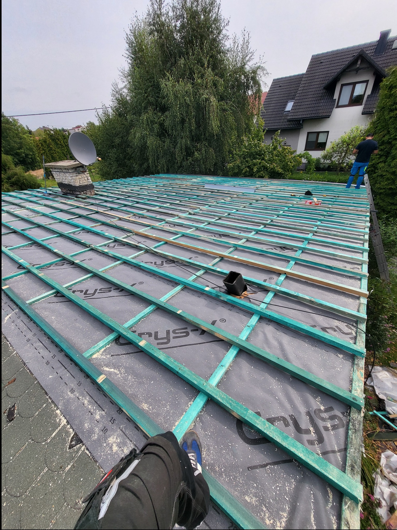 Konstrukcja dachu z łatami i membraną, widok z góry, częściowo widoczne nogi osoby pracującej na dachu, dom w tle, pochmurne niebo.