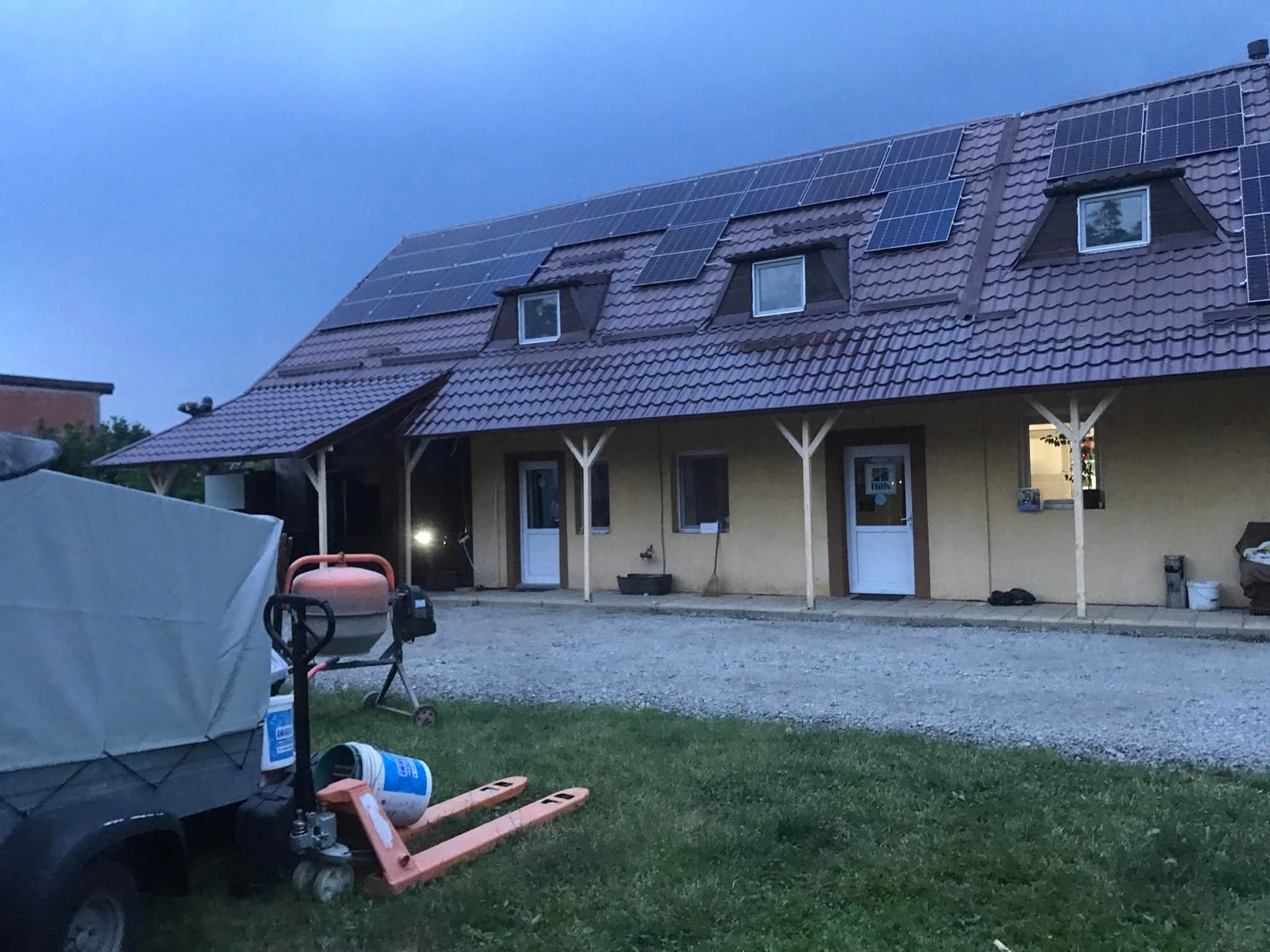 Dom z brązowym dachem i panelami słonecznymi. Na pierwszym planie przyczepa, betoniarka i paleciak. Ujęcie z dołu, w tle pochmurne niebo.