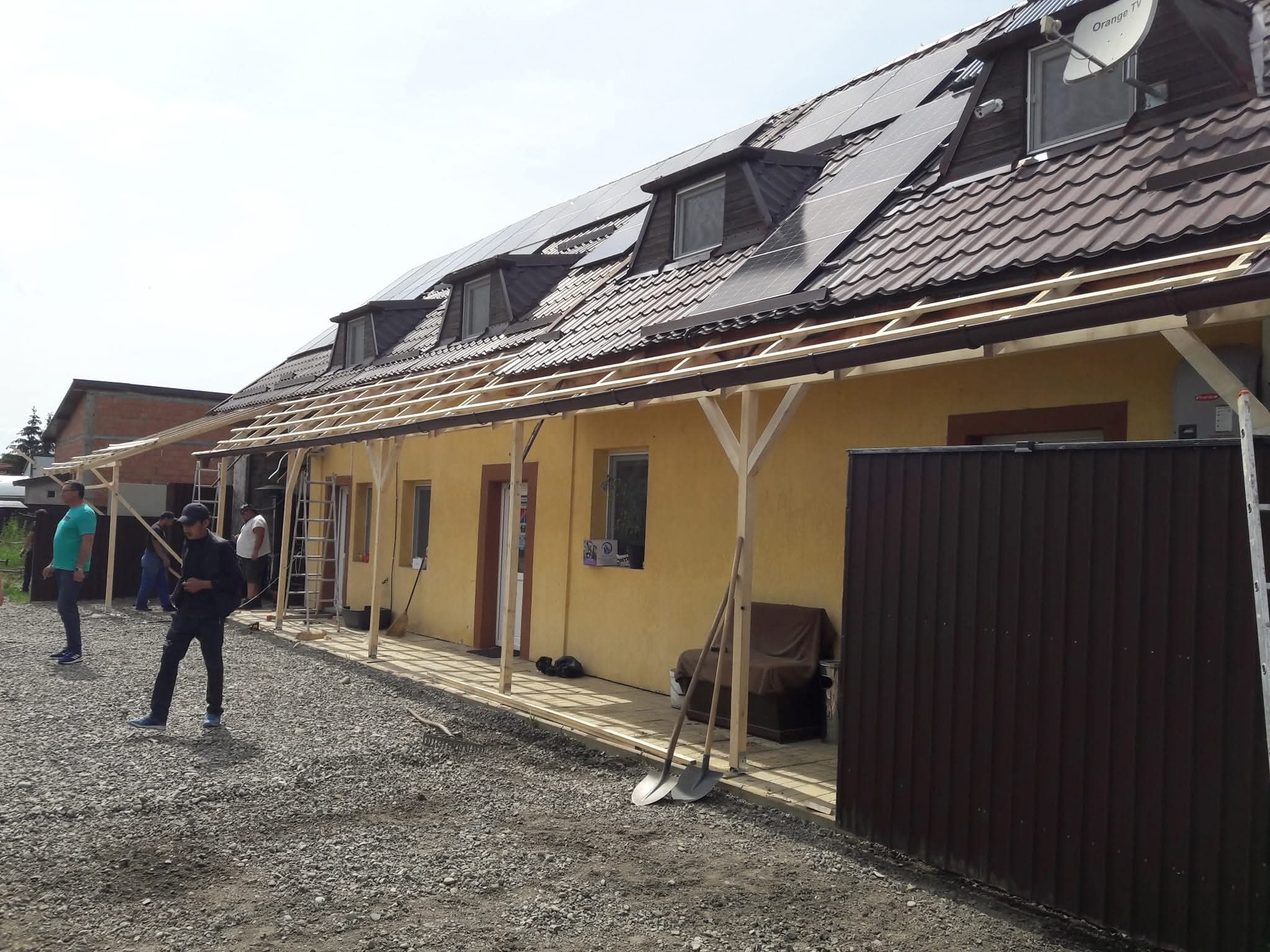 Montaż drewnianej konstrukcji zadaszenia przy żółtym budynku z brązowym dachem i panelami słonecznymi. Widoczni pracownicy, narzędzia i gruz na ziemi.
