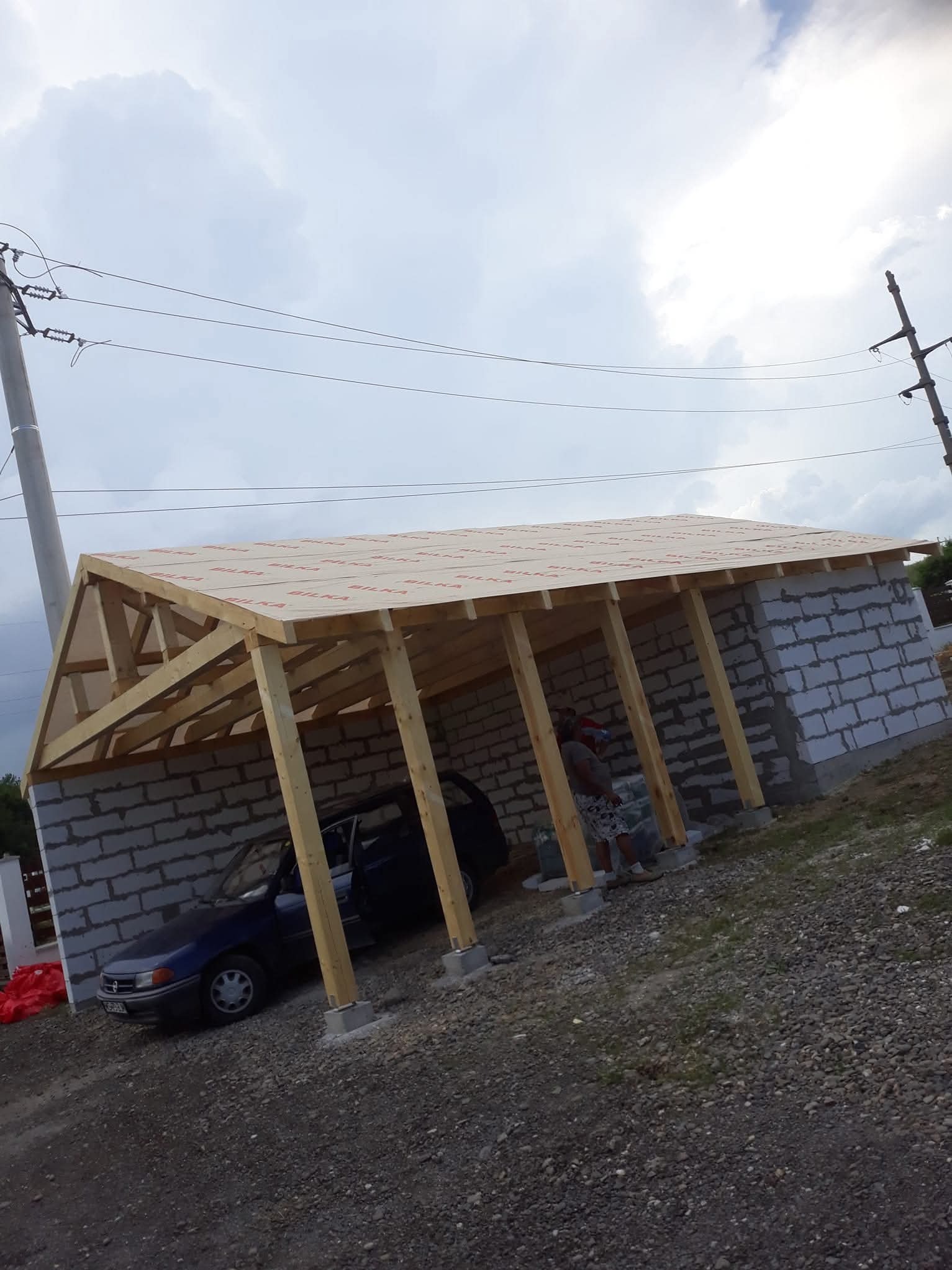 Konstrukcja dachu w trakcie budowy, widoczne bloczki betonowe, drewniane elementy więźby i auto zaparkowane wewnątrz. Częściowo pokryty dach folią.