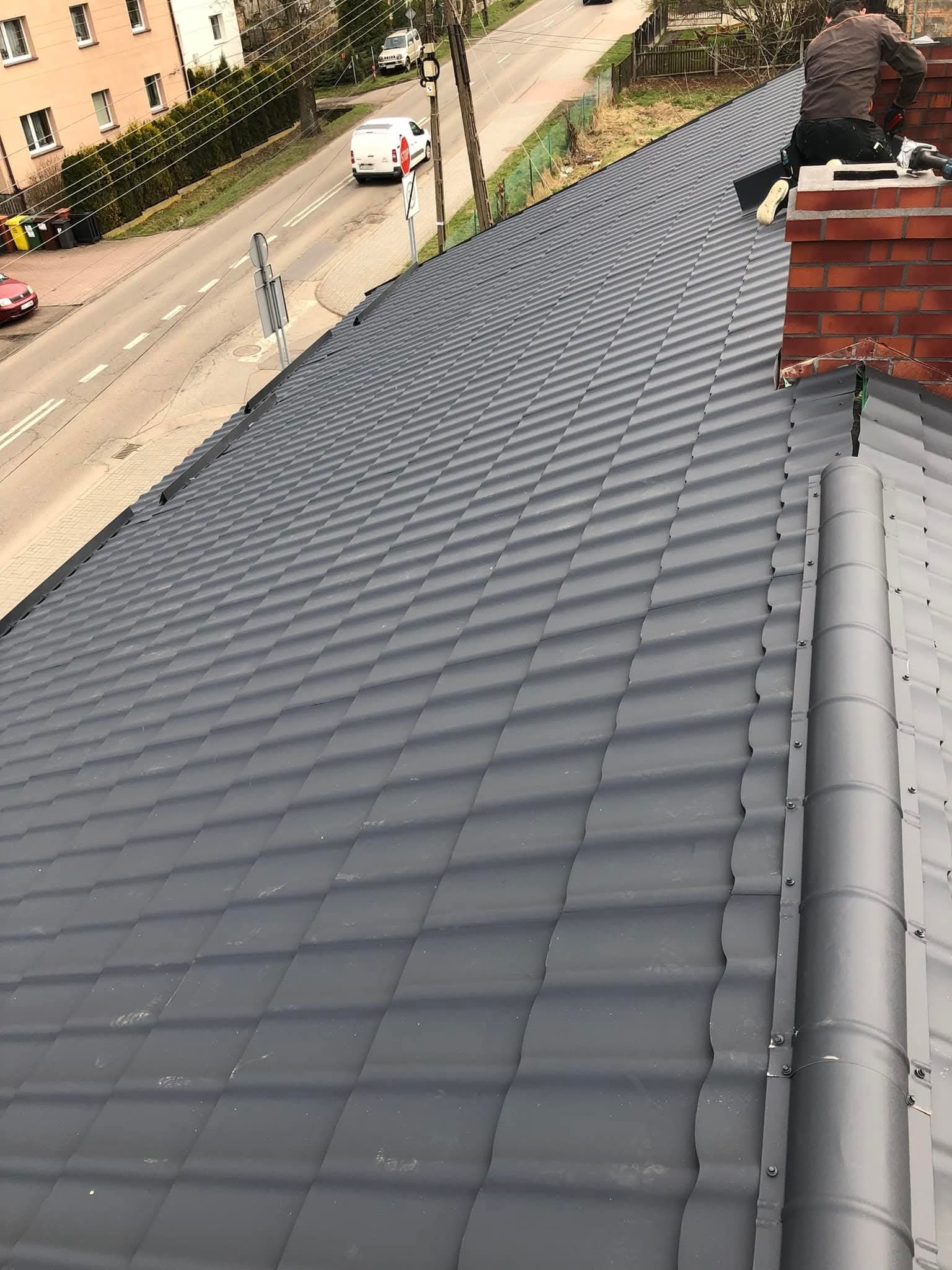 Nowy, grafitowy dach z blachy trapezowej w Katowicach. Widoczny fragment ulicy i komin. Fachowiec na dachu podczas prac dekarskich.