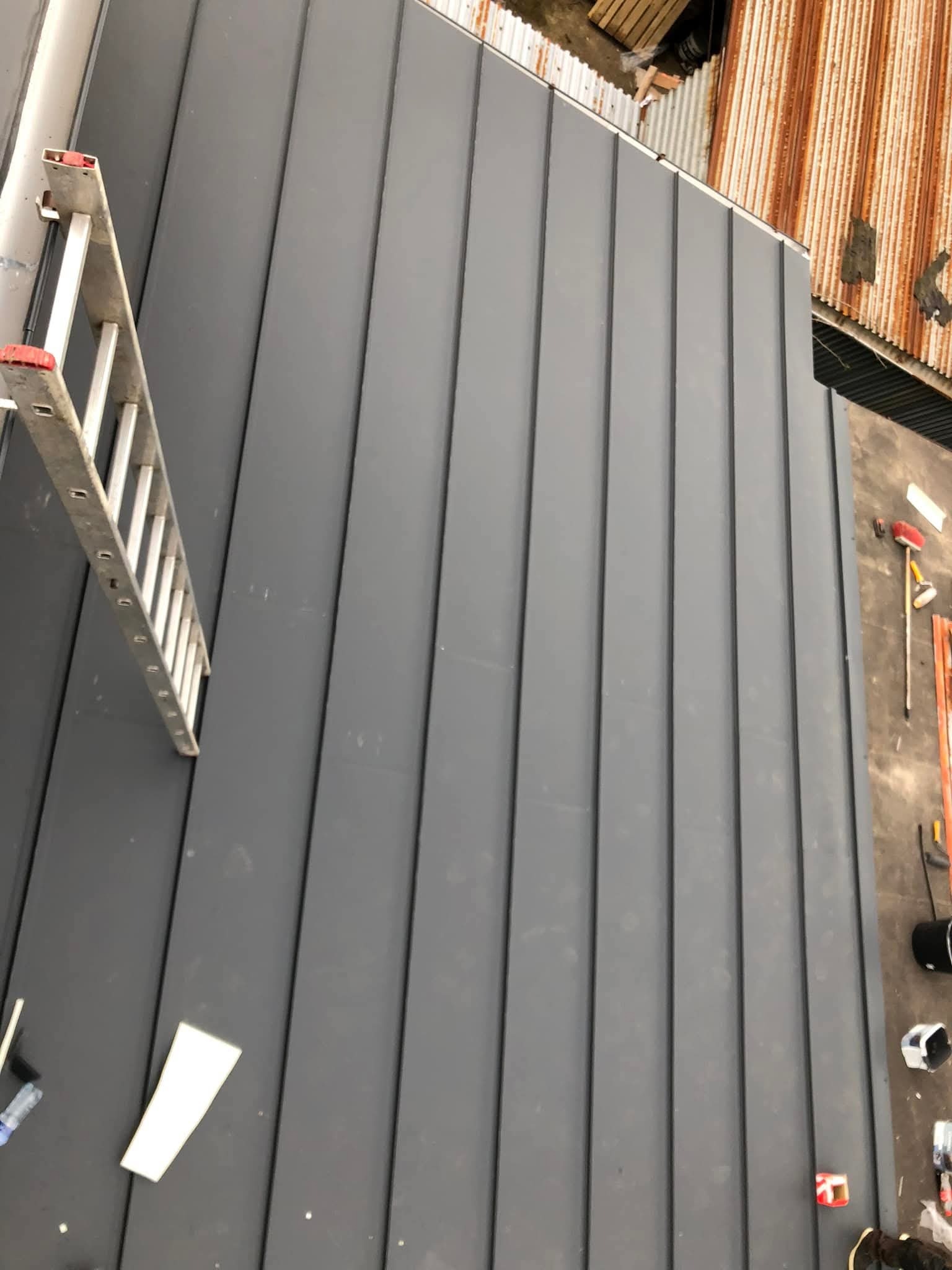 Szary dach z blachy trapezowej z drabiną opartą o jego krawędź. Widoczne narzędzia i materiały dekarskie. Dach sąsiaduje z rdzewiejącym dachem z blachy falistej.