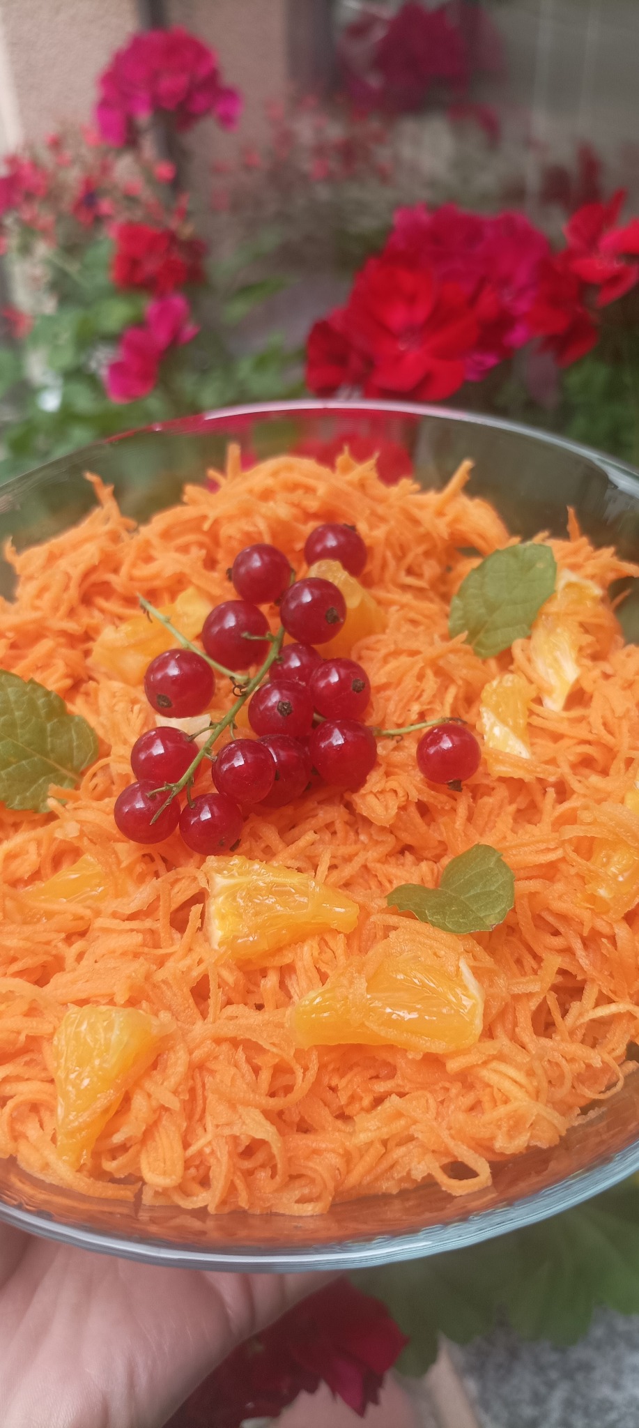 Szklana misa z marchewką julienne, kawałkami pomarańczy, czerwoną porzeczką i miętą na tle kwiatów. Orzeźwiająca, letnia sałatka owocowo-warzywna.