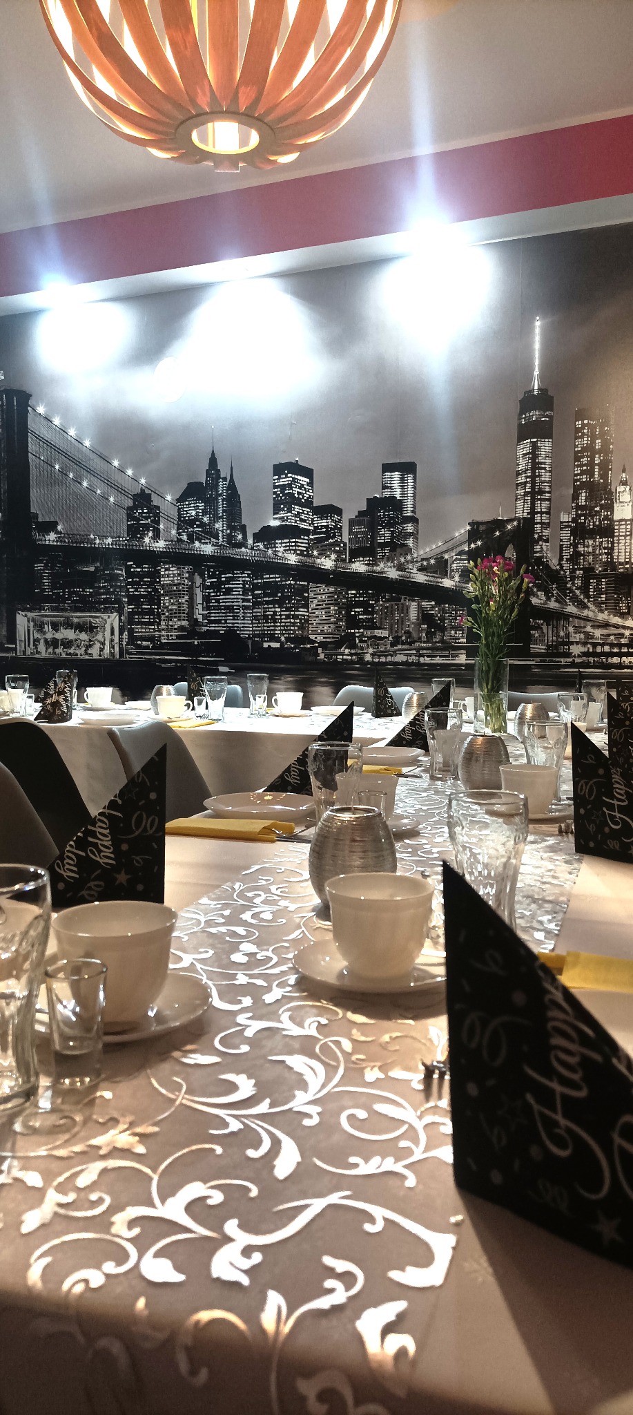 Uroczysty stół z dekoracją nowojorskiego krajobrazu w tle, nakryty białą zastawą, srebrnymi świecznikami i czarnymi czapkami urodzinowymi. Oświetlenie sufitowe.