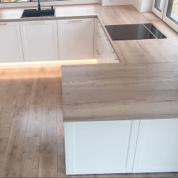 Carpintero - Nowoczesna kuchnia z białymi frontami i drewnianym blatem, oświetlenie LED pod szafkami, widok z góry, płyta indukcyjna i czarny zlew.