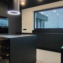 Carpintero - Nowoczesna, czarna kuchnia z wyspą i podświetlaną szafką na wino. Minimalistyczny design, okno z żaluzjami i roślina w tle. Oświetlenie LED nad wyspą.