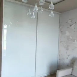 Szafa wnękowa z przesuwnymi, białymi drzwiami i delikatnym oświetleniem sufitowym w nowoczesnym wnętrzu. Widoczny fragment jasnej sofy.