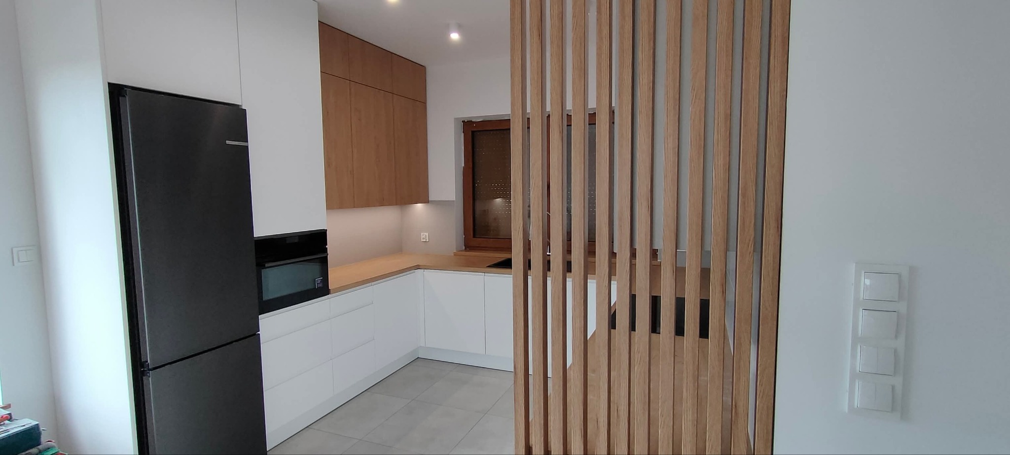 Nowoczesna kuchnia z białymi frontami i drewnianymi akcentami, czarna lodówka, drewniana ścianka działowa i szare płytki podłogowe. Oświetlenie punktowe.