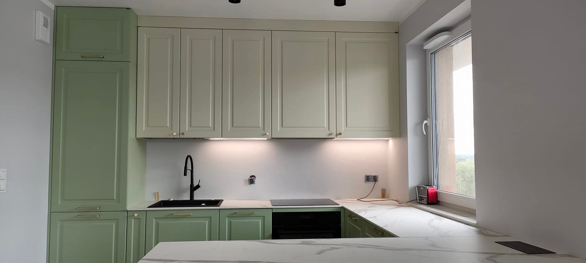 Jasna kuchnia w odcieniach zieleni i beżu, z blatem imitującym marmur, czarną armaturą i oświetleniem LED pod szafkami. Widok na okno z radiem.