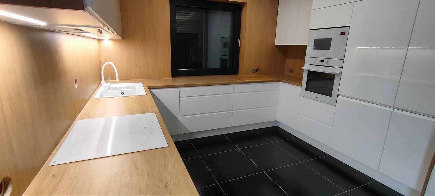 Nowoczesna kuchnia z białymi frontami i drewnianymi blatami w Wisznia Mała. Widoczny zlew, płyta indukcyjna i piekarnik w zabudowie. Czarna podłoga.