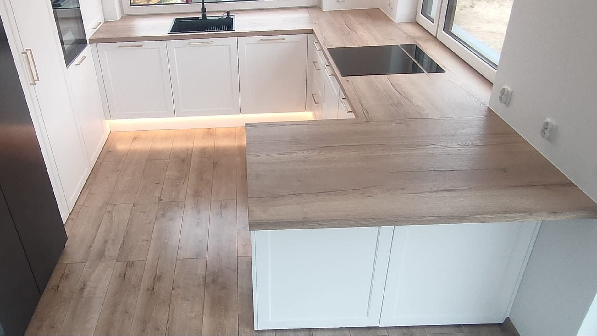 Nowoczesna kuchnia z białymi frontami i drewnianym blatem, oświetlenie LED pod szafkami, widok z góry, płyta indukcyjna i czarny zlew.