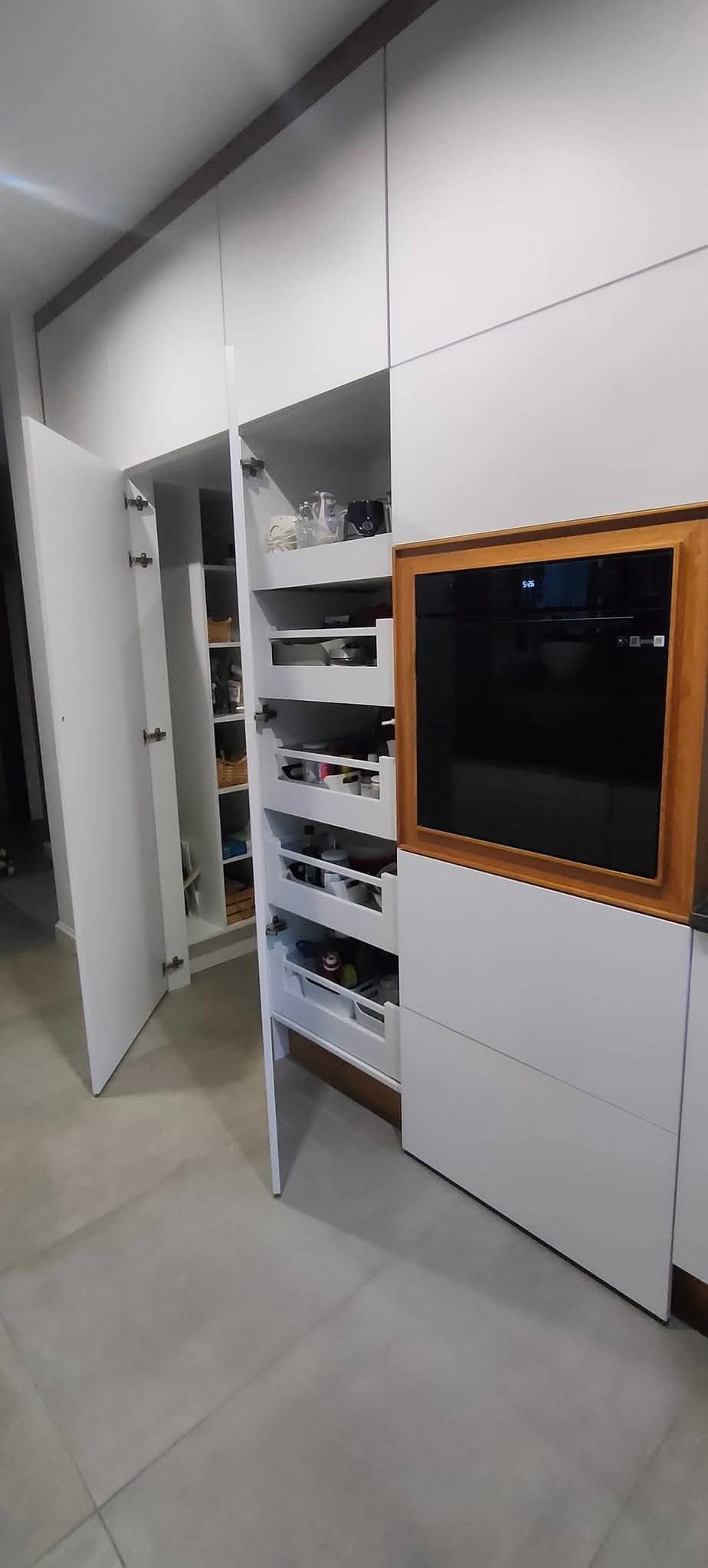 Biała kuchnia na wymiar z otwartymi szafkami i szufladami, ukazującymi zawartość. Zabudowa z piekarnikiem w drewnianej ramie. Minimalistyczny design.