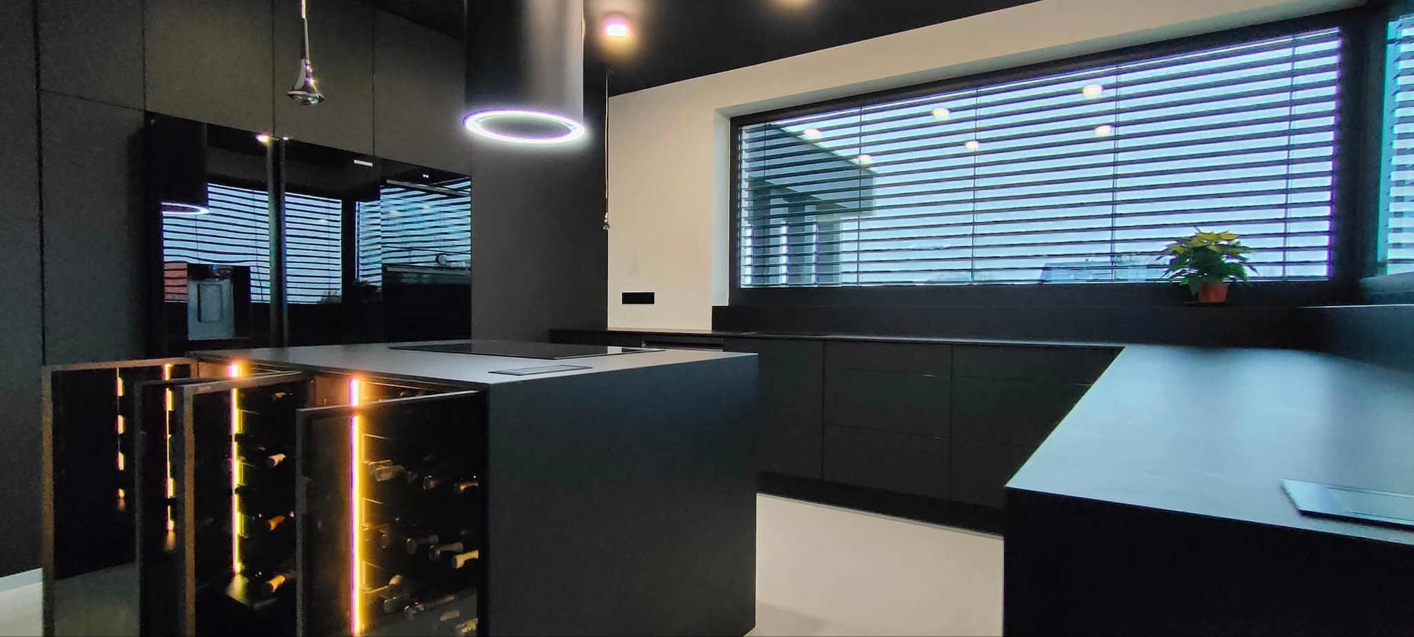 Nowoczesna, czarna kuchnia z wyspą i podświetlaną szafką na wino. Minimalistyczny design, okno z żaluzjami i roślina w tle. Oświetlenie LED nad wyspą.