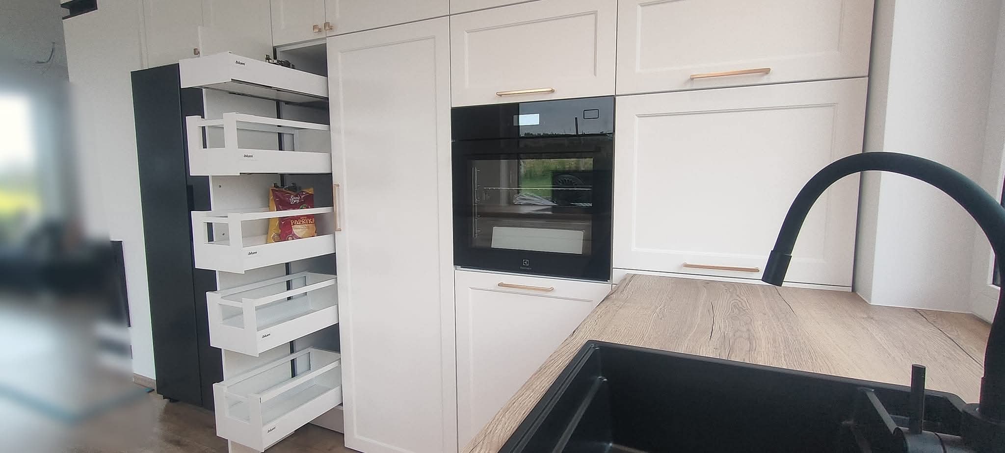 Biała kuchnia z wysuwaną szafką cargo Blum, piekarnikiem Electrolux i czarnym zlewem. Blat drewnopodobny. Ujęcie z perspektywy zlewu, widoczne detale okuć.