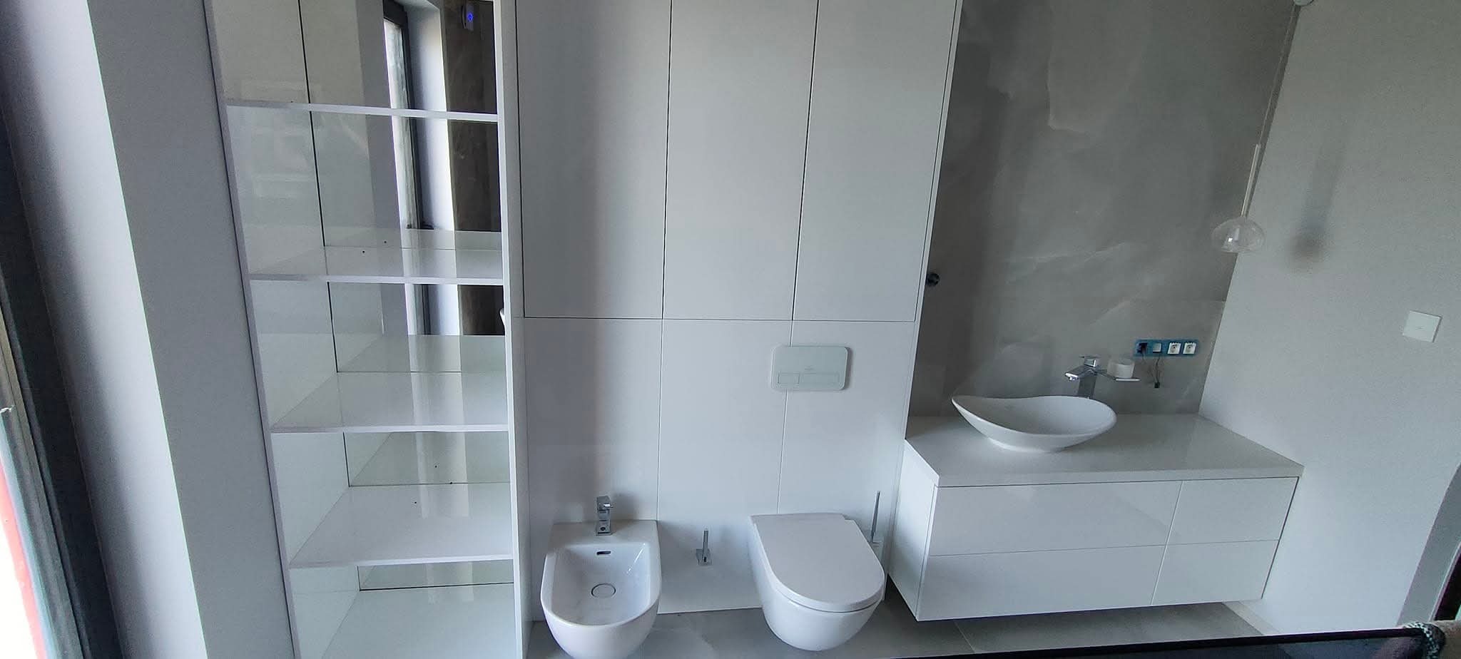 Nowoczesna, biała łazienka z szafami na wymiar, bidetem i umywalką nablatową. Minimalistyczny design, szara ściana z efektem marmuru.