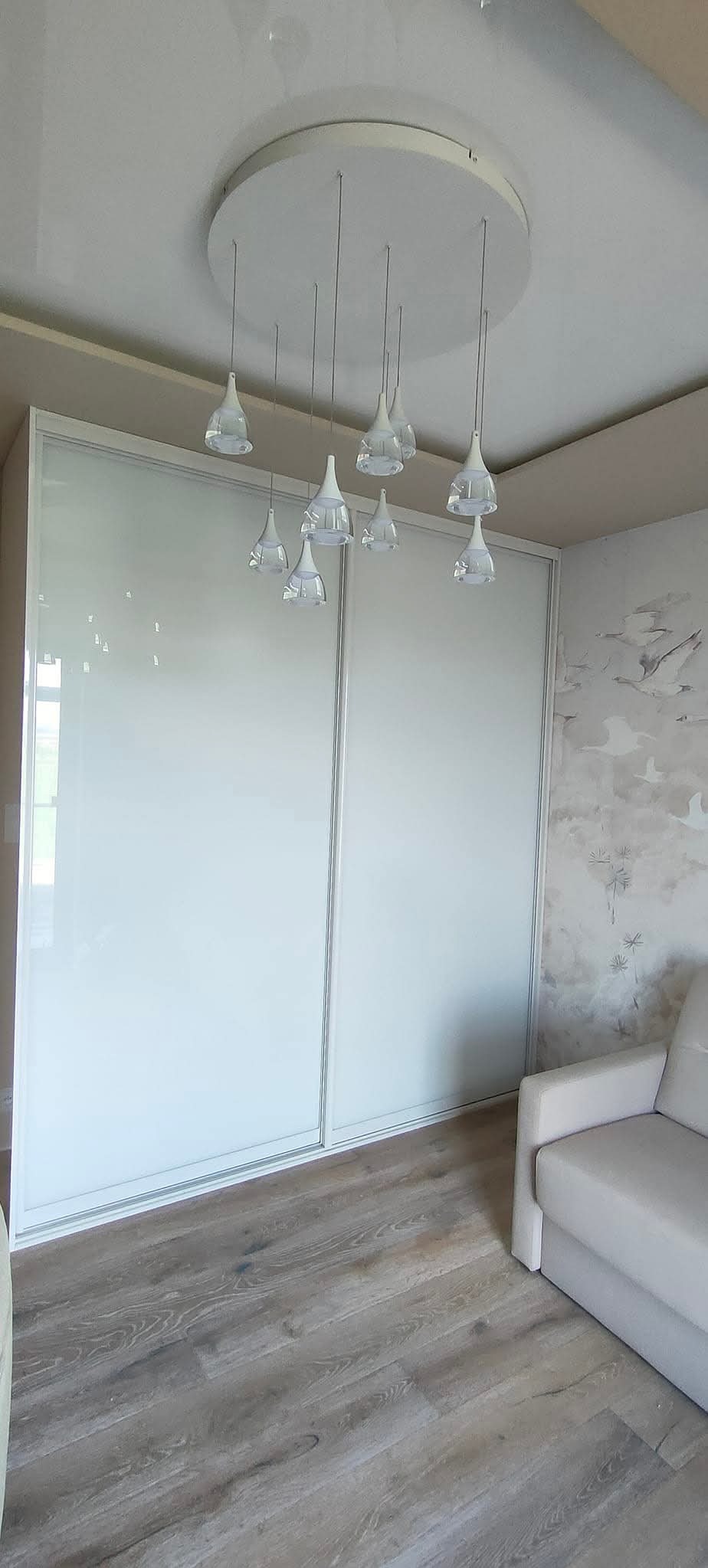 Szafa wnękowa z przesuwnymi, białymi drzwiami i delikatnym oświetleniem sufitowym w nowoczesnym wnętrzu. Widoczny fragment jasnej sofy.