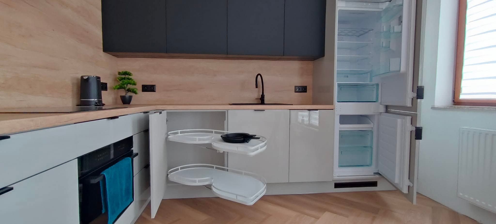 Nowoczesna kuchnia z szarymi szafkami górnymi, drewnianym blatem i otwartą szafką narożną z półkami obrotowymi, obok otwarta lodówka.