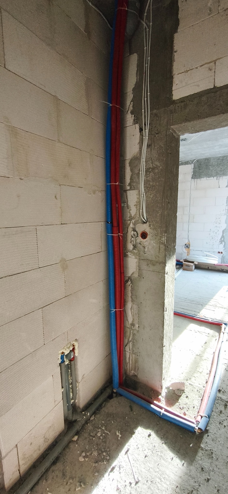 Instalacja wodna w budynku w trakcie budowy. Czerwone i niebieskie rury prowadzone wzdłuż ściany z betonu komórkowego, zabezpieczone opaskami. Widoczne także szare rury kanalizacyjne.